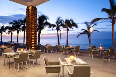Outdoor restaurant by the ocean at sunset, with palm trees, neatly set tables, and lit torches creating a serene ambiance.