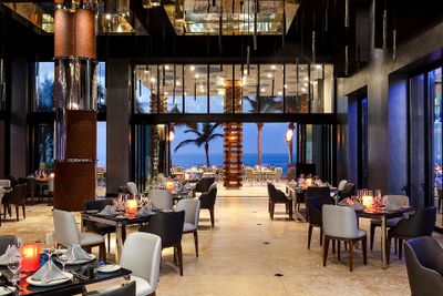Elegant restaurant interior with modern decor, set tables, and a view of palm trees and the ocean through large windows.