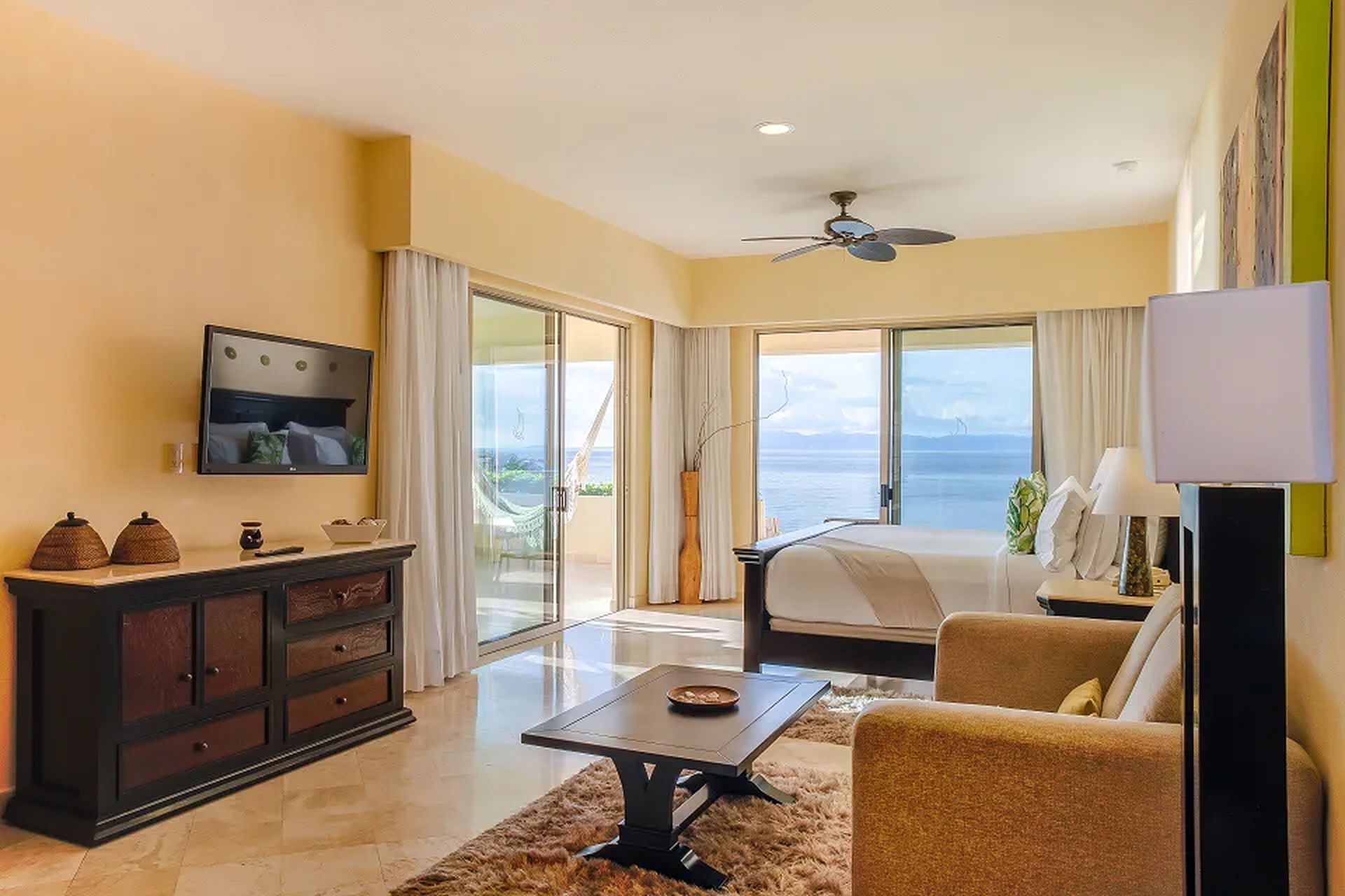 Spacious hotel room with a bed, sofa, and TV, featuring large windows with a view of the ocean and a balcony.