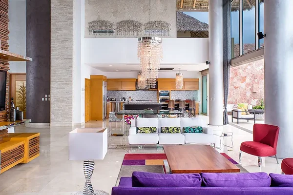 Spacious modern living room with colorful furniture, large chandeliers, and an open kitchen. Floor-to-ceiling windows offer natural light.