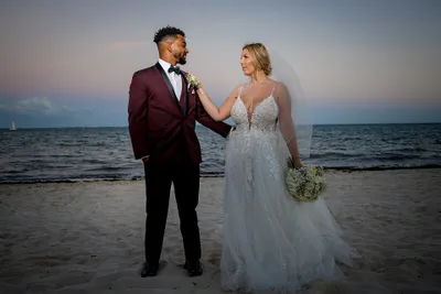 A bride and groom stand on a sandy beach at sunset, gazing at each other lovingly, with the ocean in the background.