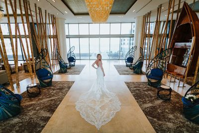 Bride in a white lace gown stands in a spacious, elegant room with tall bamboo decor, blue chairs, and large windows.