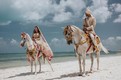 A couple in traditional attire rides ornately decorated horses on a sandy beach under a clear blue sky.