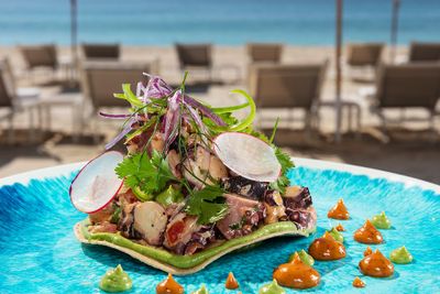 A vibrant seafood salad with octopus, radish, and cilantro on a blue plate, garnished with colorful sauces, set against a beach backdrop.