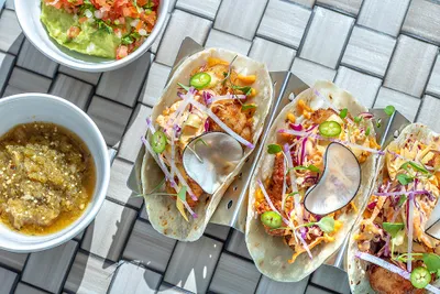Three tacos with toppings on metal holders, accompanied by bowls of salsa and guacamole, on a woven surface.
