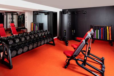 Modern gym with red equipment, dumbbells, colorful kettlebells, and a black wall. Orange flooring adds contrast to the sleek design.