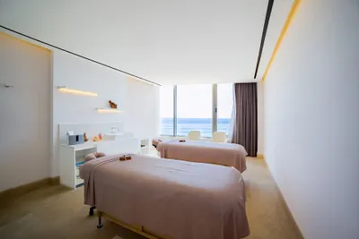 Minimalist spa room with two massage tables, soft lighting, and a large window offering an ocean view.