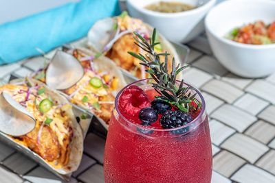 A berry cocktail garnished with rosemary and berries, with tacos and side dishes in the background on a tiled table.