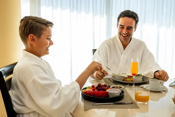 A child and an adult in white robes enjoy a breakfast with fruit, juice, and pastries at a table by a window. Both are smiling.