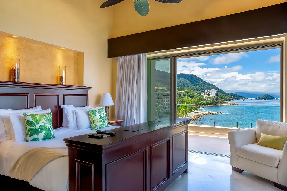 Luxurious beachfront hotel room with tropical decor, large bed, and ocean-view balcony overlooking lush greenery and the sea in Cancun.