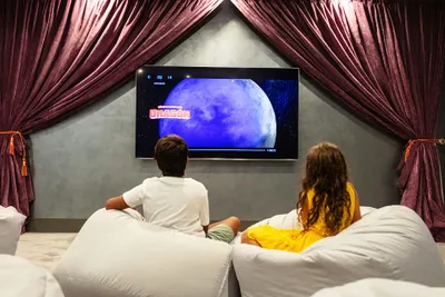 Two children sit on bean bags in a cozy room with purple curtains, watching a movie on a large screen showing a moonlit scene.