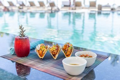 Tacos and sauces on a mat by a pool, with a red drink garnished with rosemary, and sun loungers in the background.