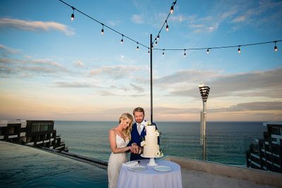 A couple in wedding attire cuts a white cake on a rooftop overlooking the ocean at sunset, with string lights above.