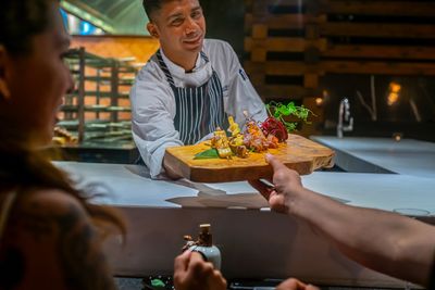 Chef presenting a wooden platter with skewered food to a customer at a counter, with a kitchen background.
