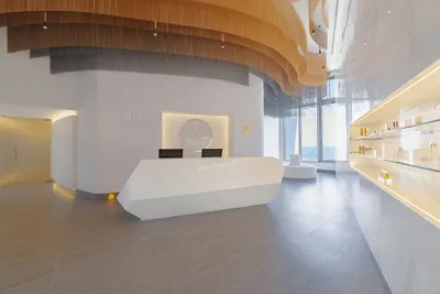 Modern spa reception with a sleek white desk, curved wooden ceiling accents, wall display of products, and large windows with ocean view.