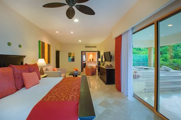 Spacious hotel room with a king-size bed, sitting area, TV, and sliding glass doors leading to a balcony with outdoor seating and lush greenery.