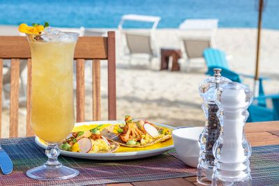 A tropical drink and a plate of food on a table by the beach, with lounge chairs and the ocean in the background.