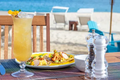 A tropical drink and a plate of food on a table by the beach, with lounge chairs and the ocean in the background.