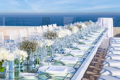 Elegant outdoor dining setup with a long table, white chairs, glassware, and white floral centerpieces, overlooking a clear blue ocean.