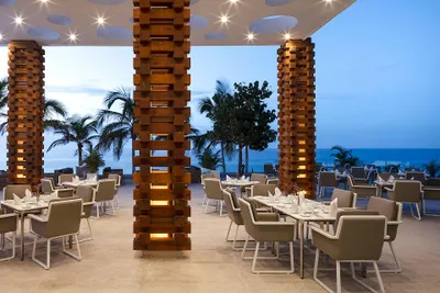 Outdoor restaurant with modern wooden columns, elegant tables, and chairs. Overlooking the ocean, surrounded by palm trees at dusk.