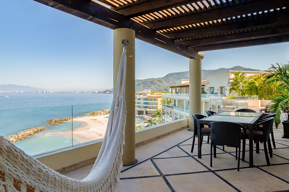 Beachfront terrace with a hammock and dining set overlooking turquoise waters and sandy beach, framed by lush greenery and distant mountains.
