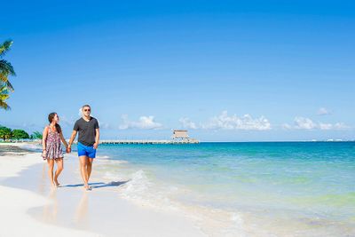 Couple holding hands, walking along a pristine, sunny beach with clear turquoise water and a wooden pier in the distance.