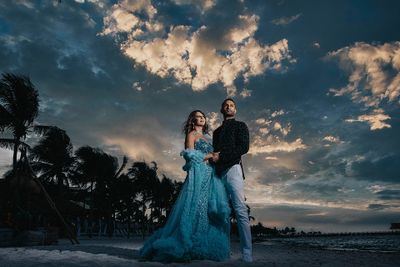 A couple stands elegantly on a beach at sunset, with dramatic clouds and palm trees in the background.