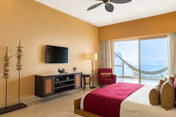 Modern hotel room with a large bed, red accents, a wall-mounted TV, and a hammock on the balcony overlooking the ocean.