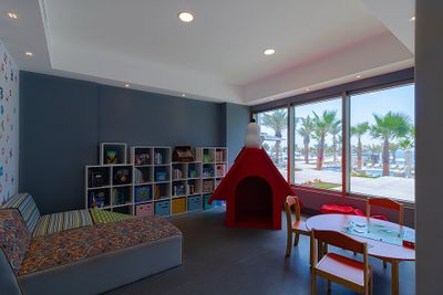 Bright children's playroom with colorful furniture, bookshelves, a small table with chairs, and a window view of palm trees and the sea.