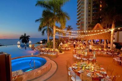 Outdoor dining area by a pool at sunset, with string lights, elegantly set tables, palm trees, and a view of the ocean and sky.