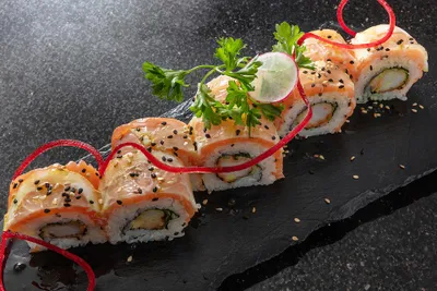 Sushi rolls topped with salmon and black sesame seeds, garnished with parsley, radish, and red decorative strands on a black slate.