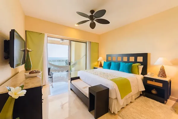 Bright hotel room with a king bed, green accents, wall-mounted TV, and balcony with ocean view. A hammock is visible outside the sliding doors.