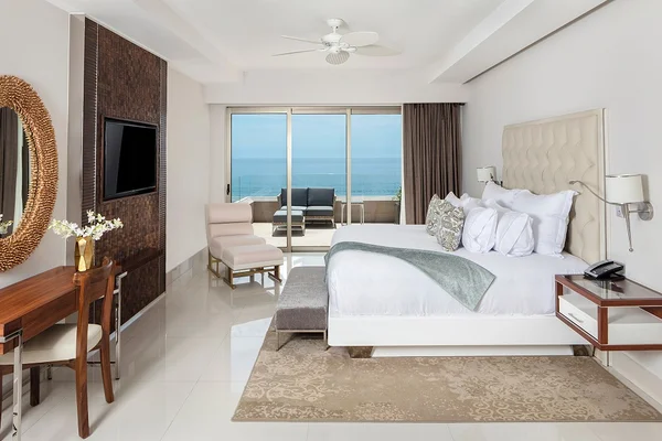 Elegant hotel room with a large bed, ocean view balcony, chairs, desk, TV, and neutral decor.