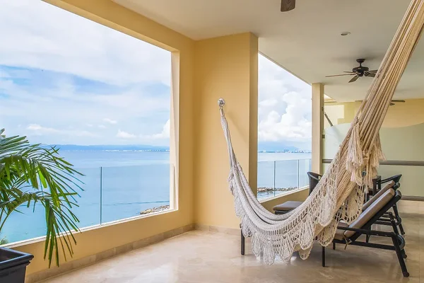 A balcony with a hammock and lounge chairs overlooks a calm ocean under a partly cloudy sky, framed by a large open window.