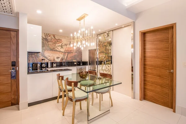 Modern dining area with glass table, wooden chairs, and a chandelier. Mosaic wall art and wooden doors enhance the elegant ambiance.