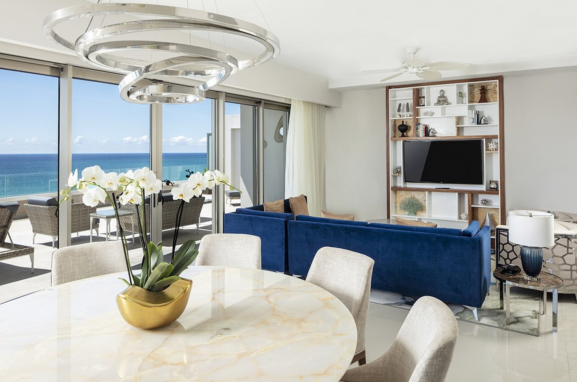 Modern living room with ocean view, featuring a marble dining table, blue sofa, large windows, and a wall-mounted TV. Elegant decor and lighting.