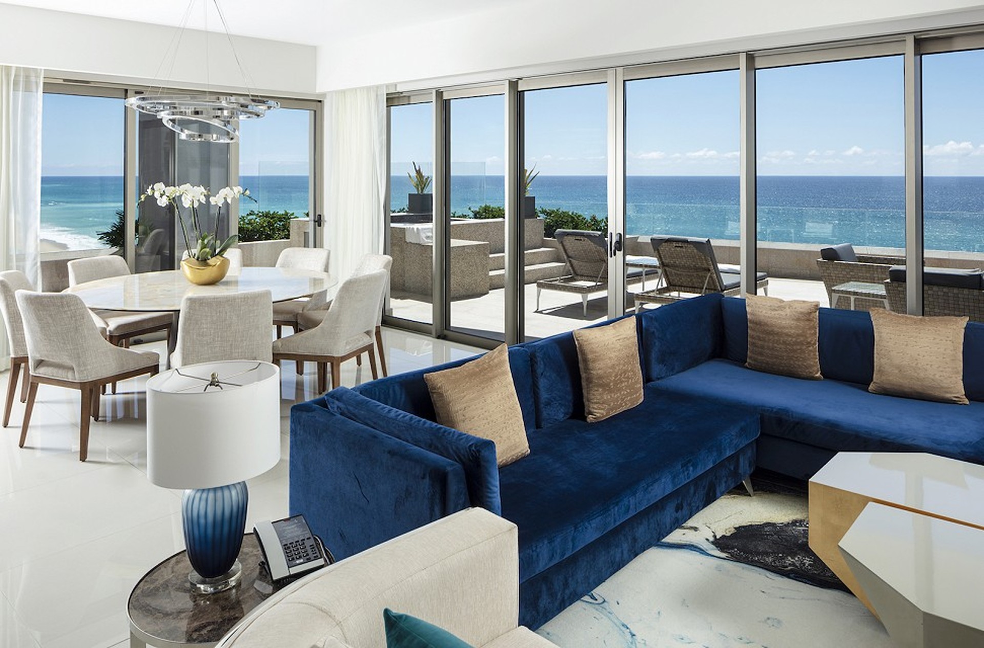 Luxurious living room with a blue sectional sofa, glass doors opening to an ocean-view balcony, and a dining area with a round table and chairs.