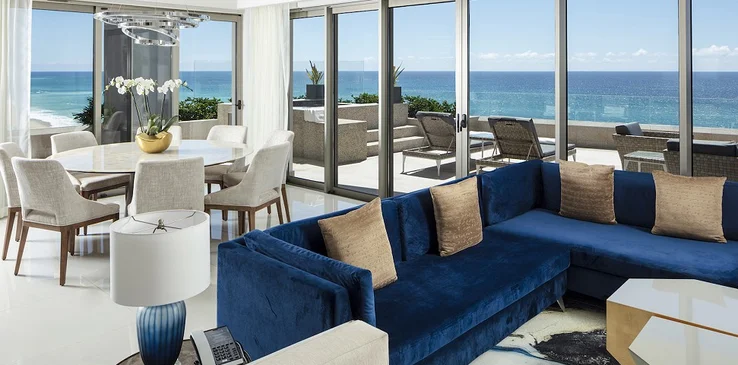 Luxurious living room with a blue sectional sofa, glass doors opening to an ocean-view balcony, and a dining area with a round table and chairs.
