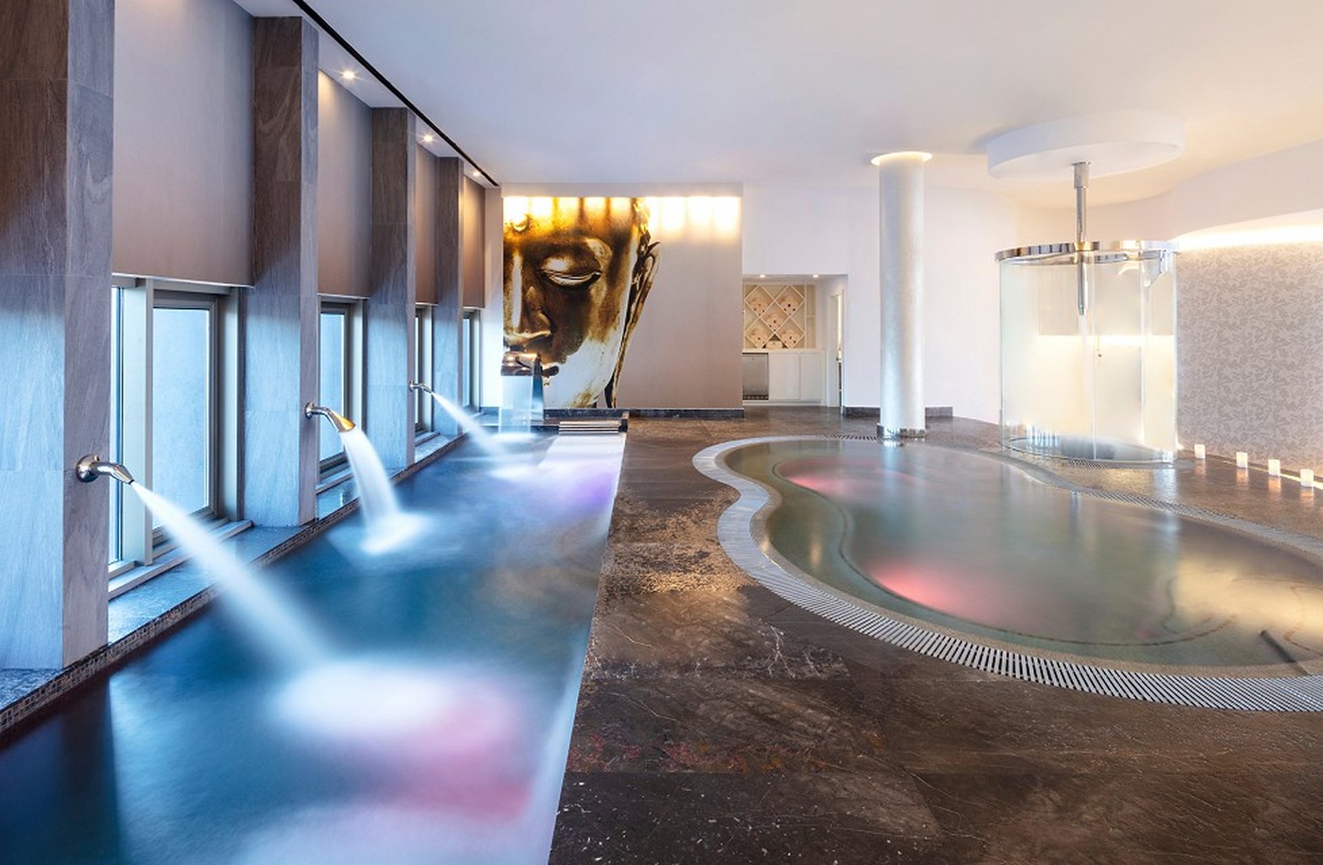 Luxurious indoor spa with a modern pool, water jets, and a large Buddha face artwork on the wall, creating a serene atmosphere.