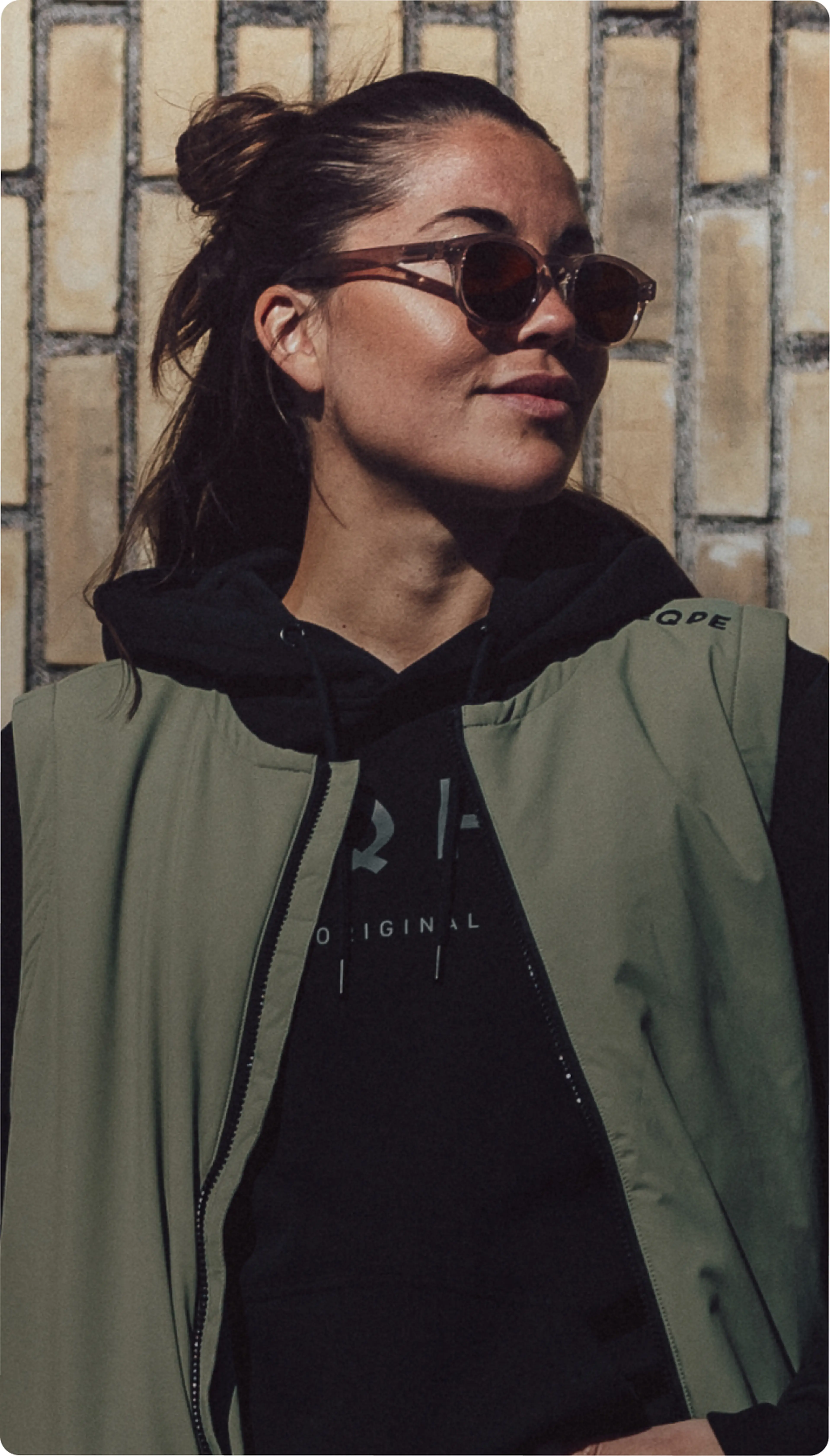 A person with sunglasses, wearing a black hoodie and green vest, stands in front of a brick wall, looking off to the side.