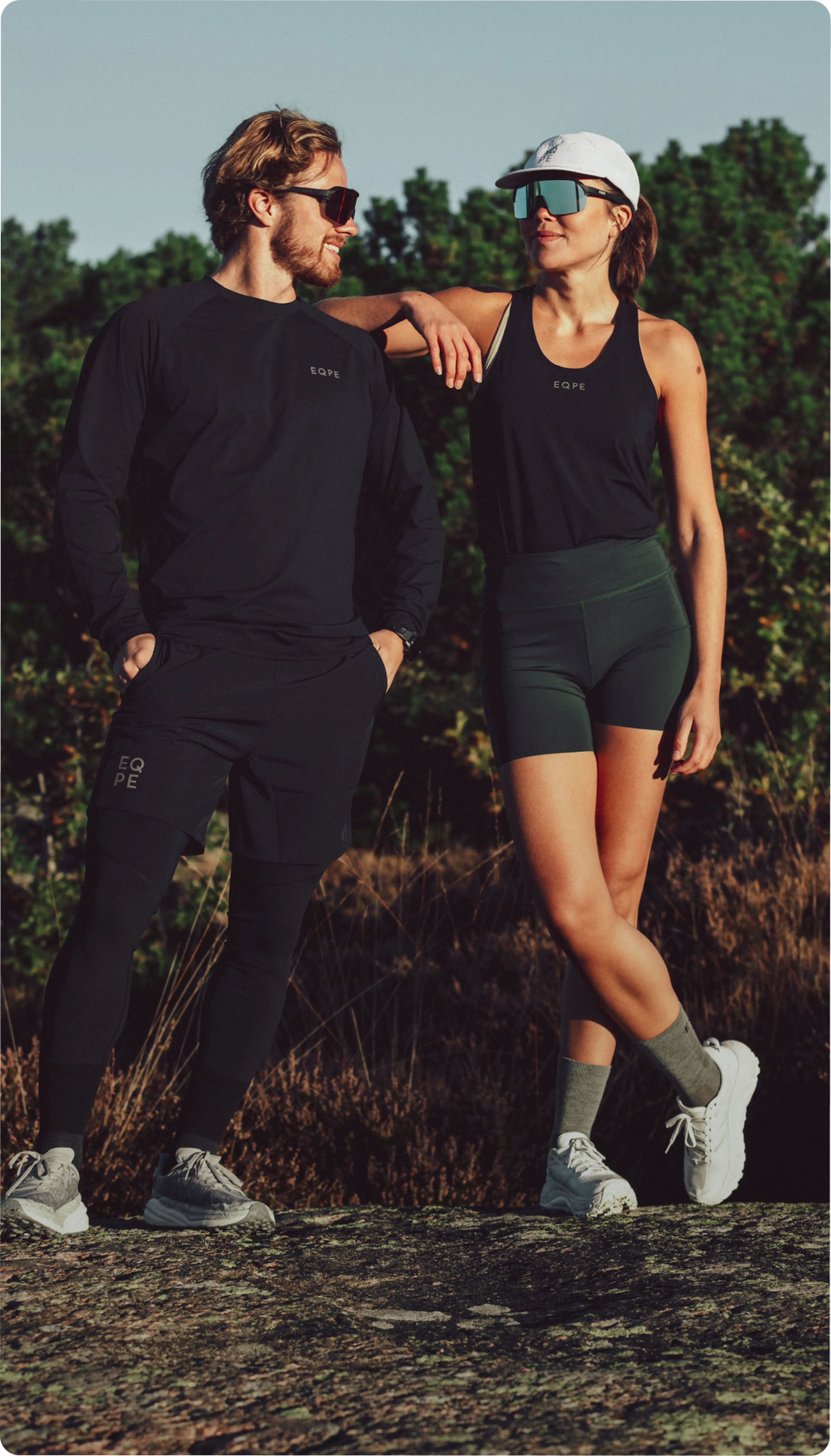 A man and woman wearing sporty black outfits, sunglasses, and sneakers, standing outdoors on a sunny day, smiling at each other.