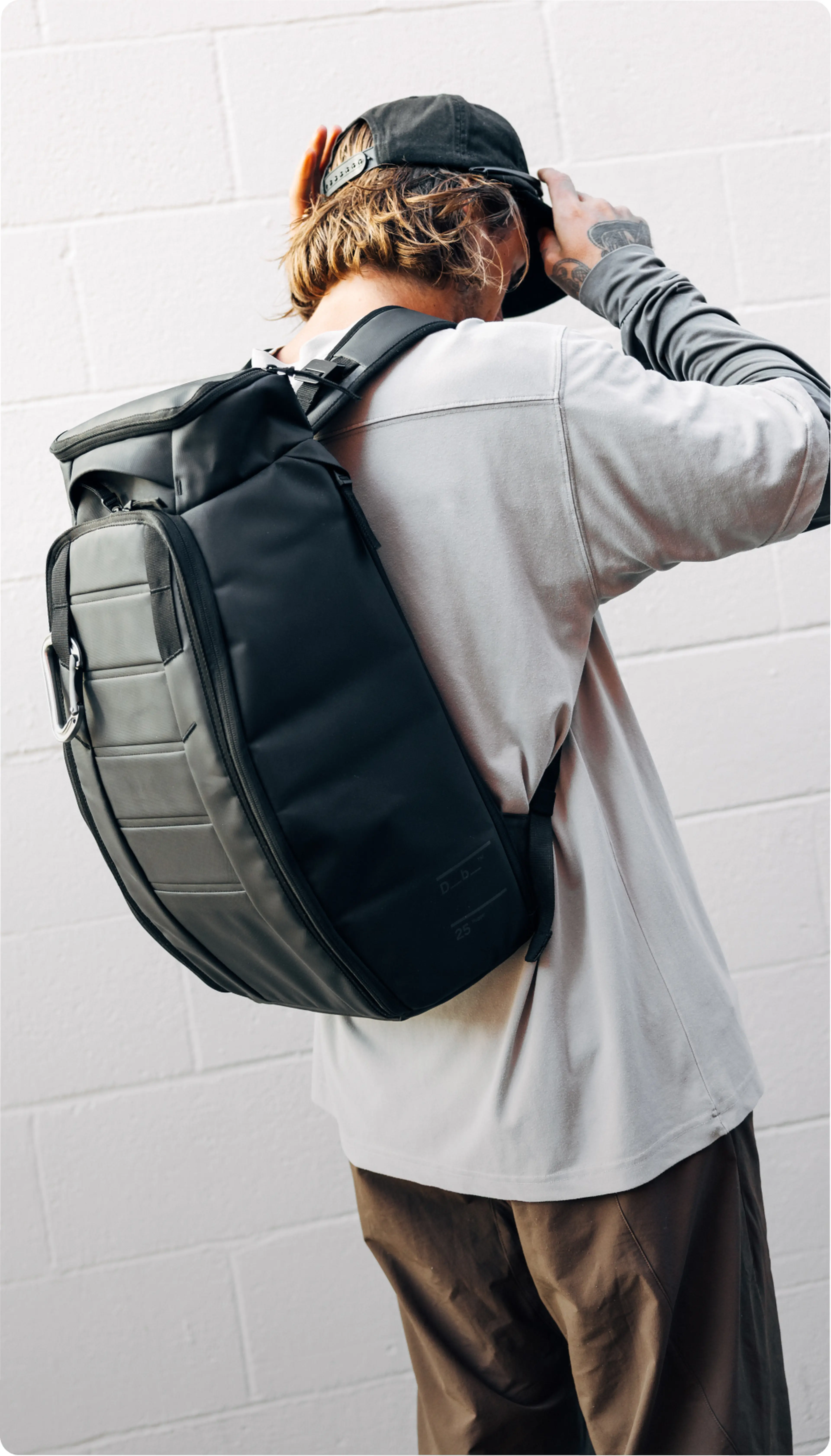 Person facing away, wearing a black backpack and a baseball cap, adjusting the cap with one hand against a white brick wall.