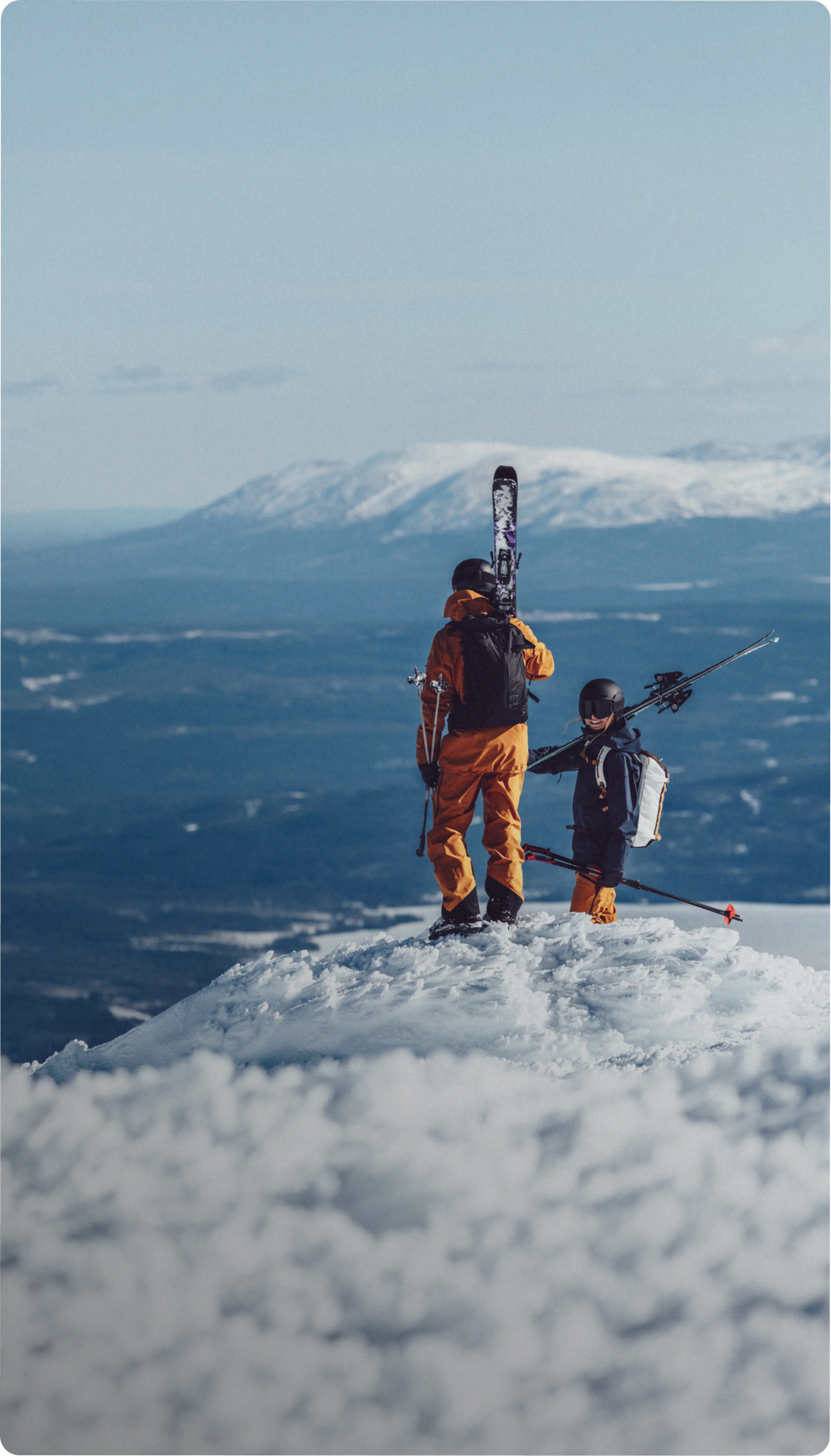 Two skiers in bright outfits standing on a snowy peak, overlooking distant mountains and valleys under a clear blue sky.