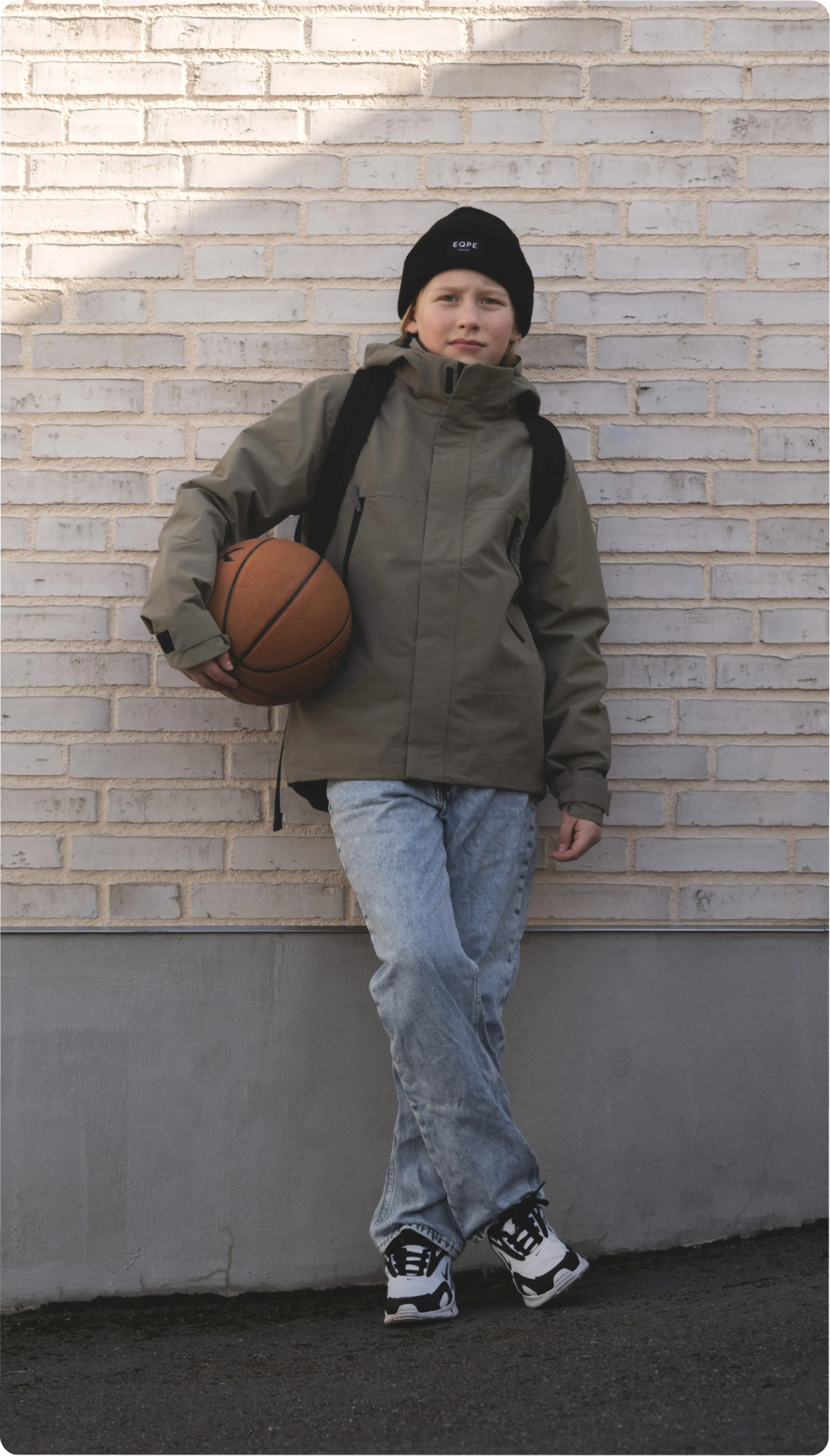 A person in a green jacket and black beanie leans against a brick wall, holding a basketball, wearing jeans and sneakers.