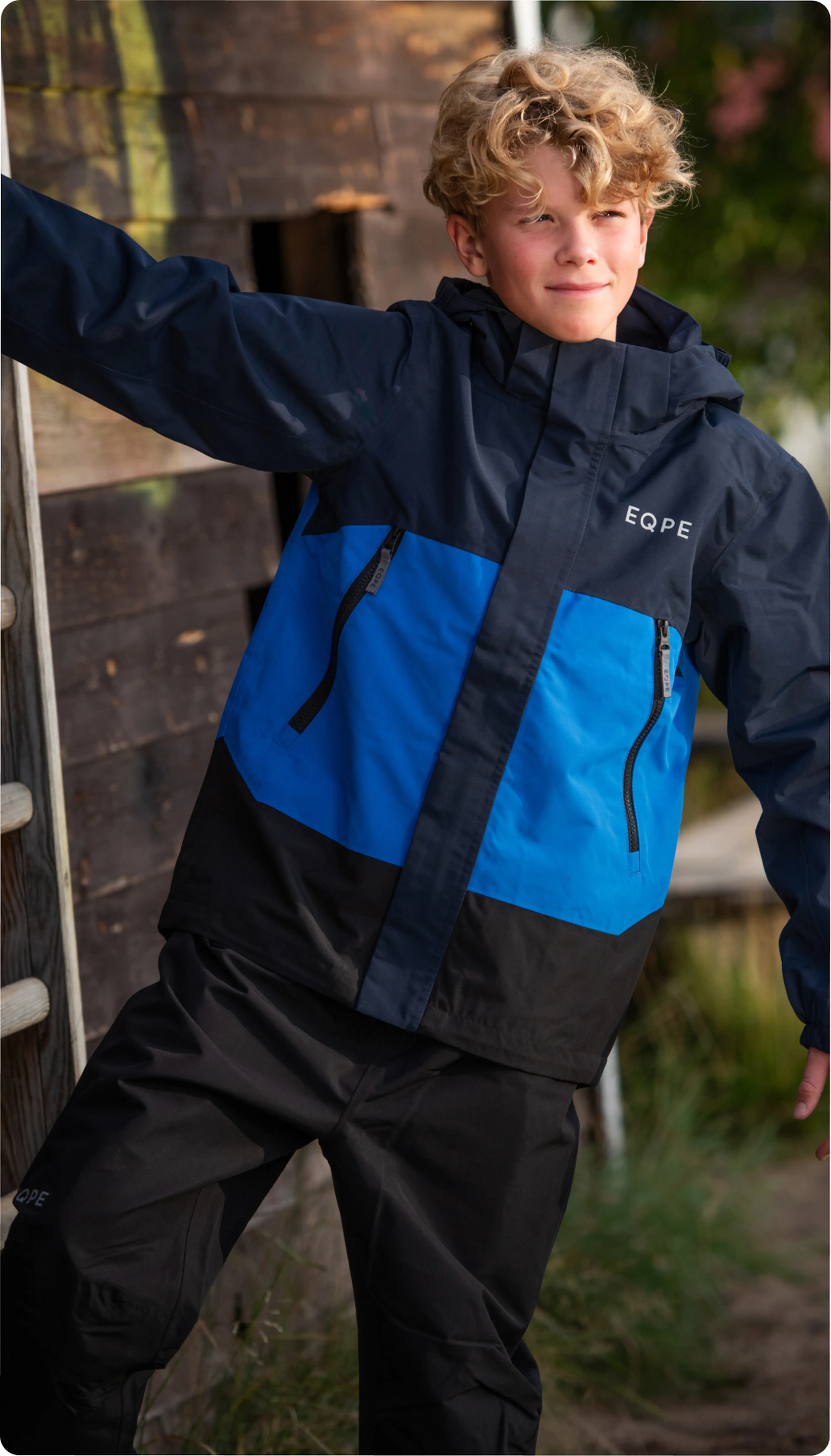 A boy with curly hair wearing a blue and black jacket stands outdoors, leaning against a wooden structure, smiling slightly.