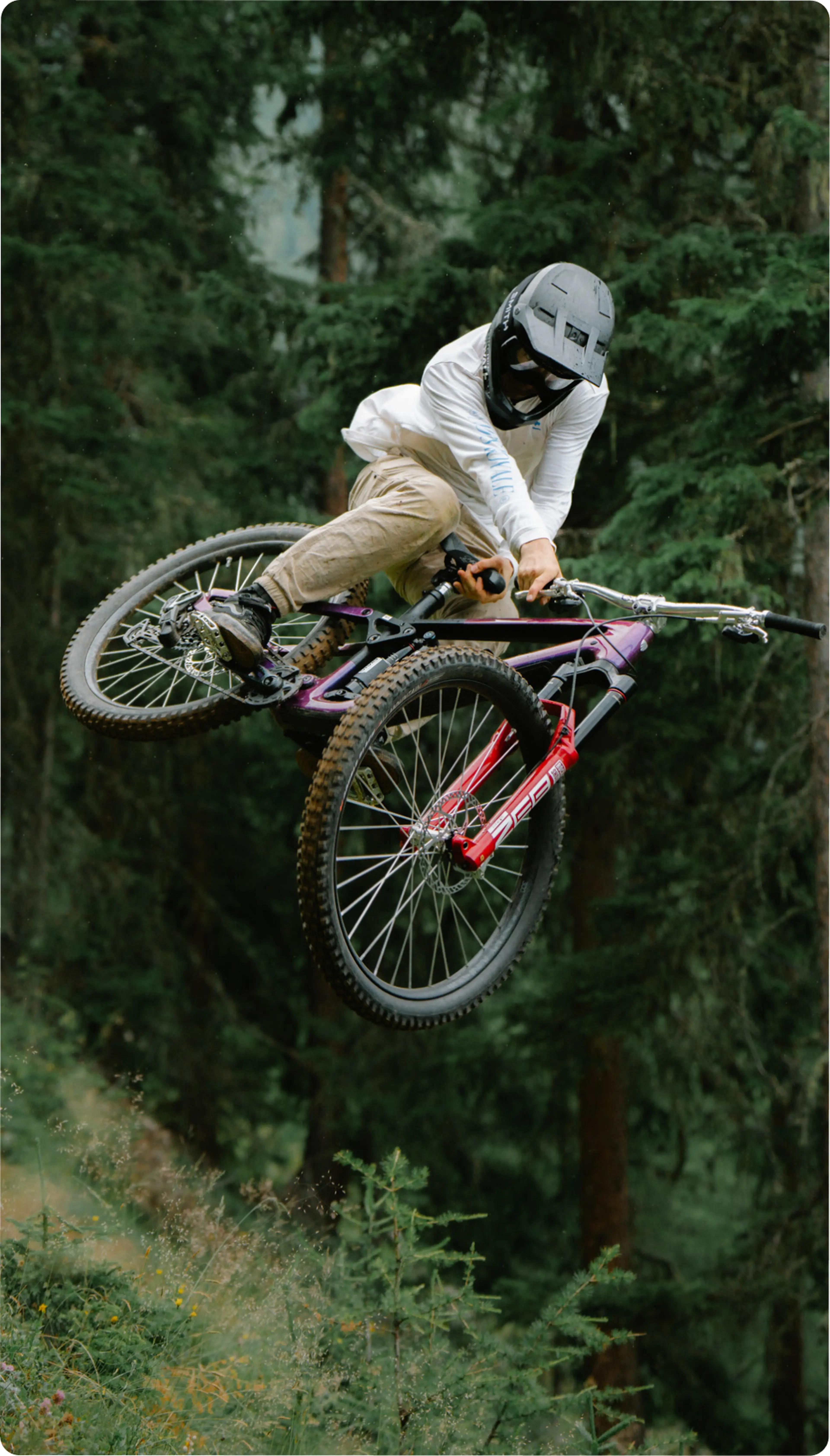A mountain biker in a helmet and gear performs a mid-air trick on a trail surrounded by dense forest greenery.
