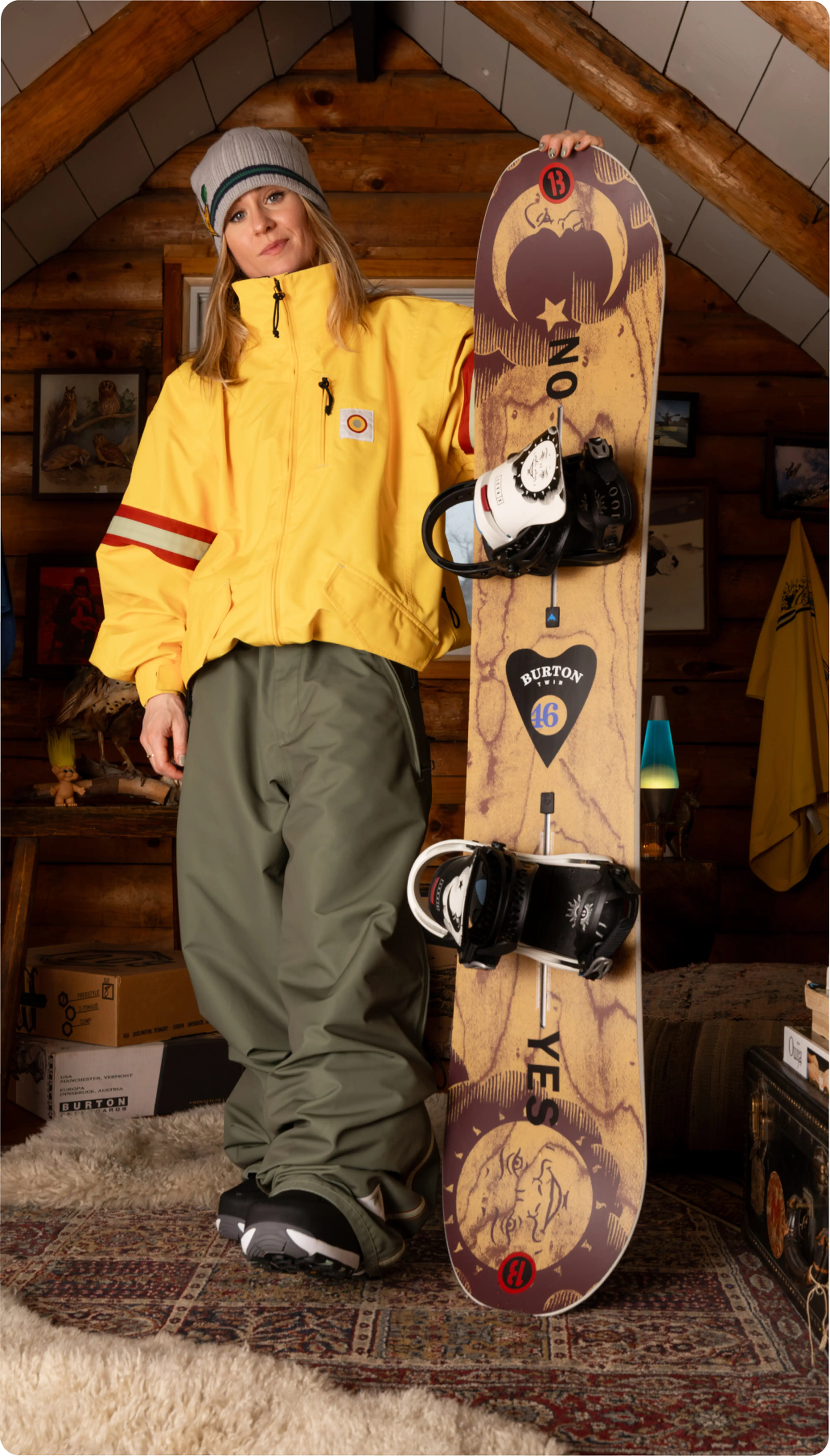 Person in a yellow jacket and gray beanie standing indoors, leaning against a snowboard with a sun face design, on a patterned rug.