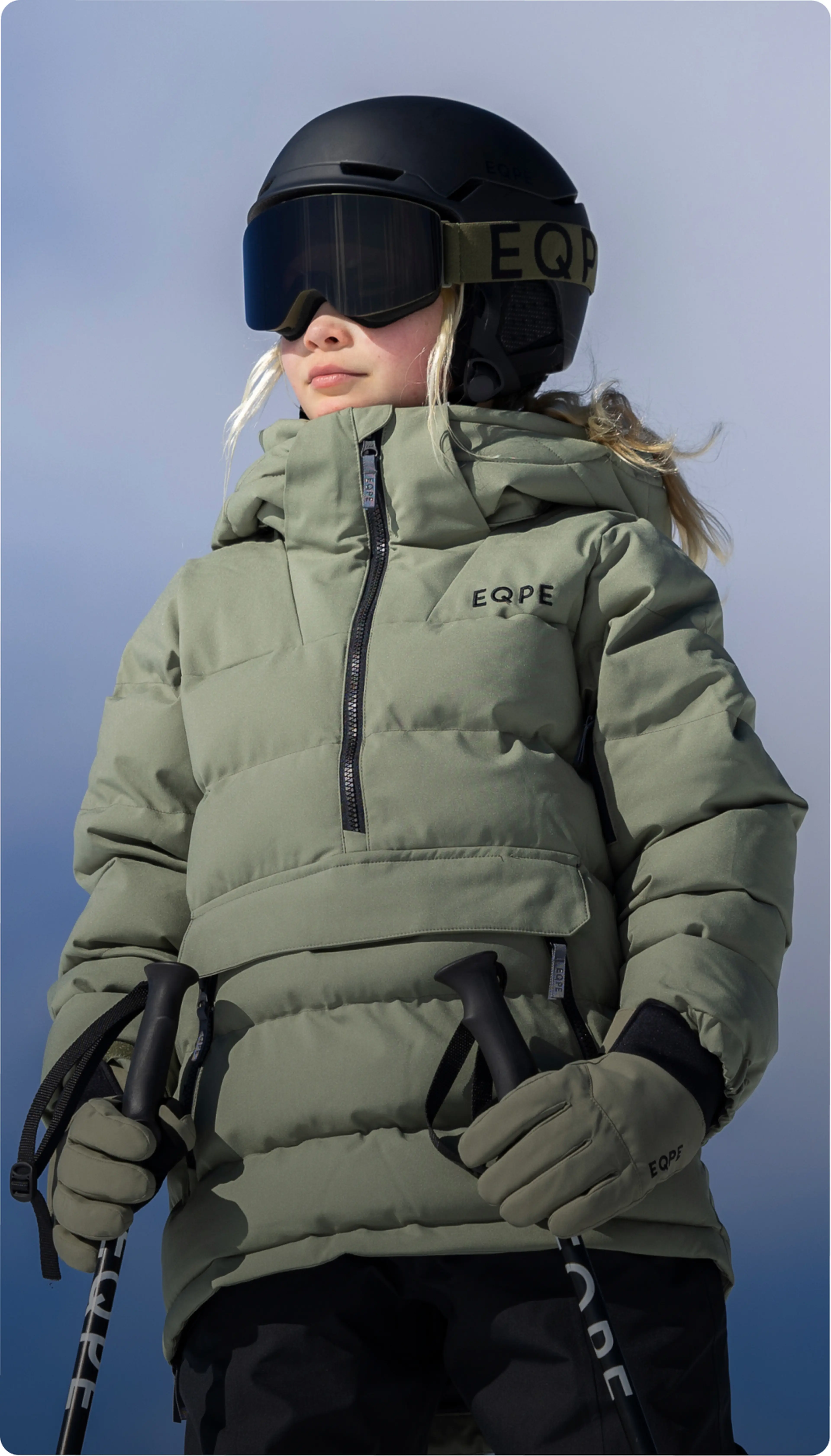 Person in green ski jacket and black helmet holding ski poles, looking up against a snowy backdrop.