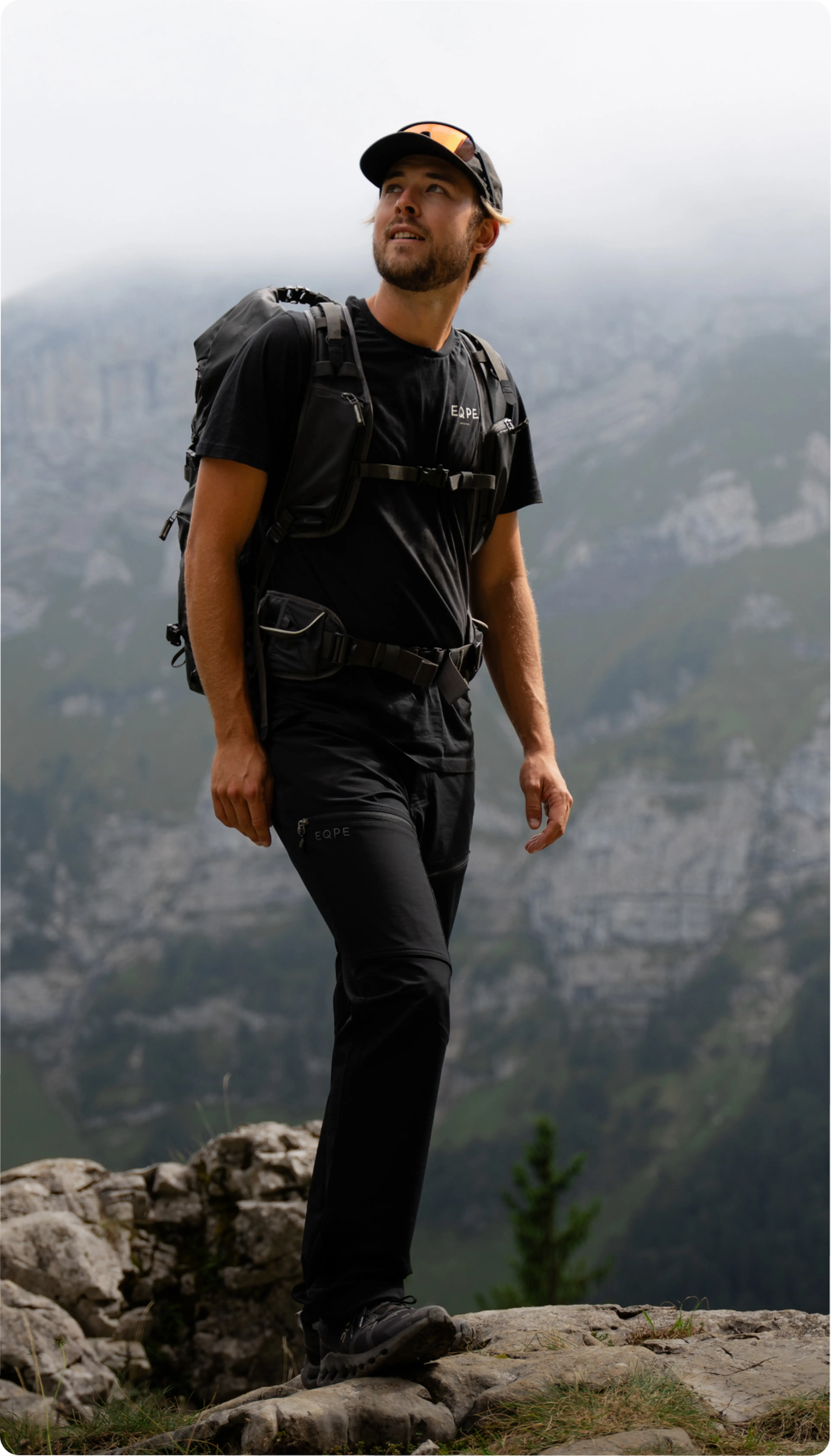 Hiker in black outfit and cap stands on a rocky ledge, gazing up, with misty mountains in the background.