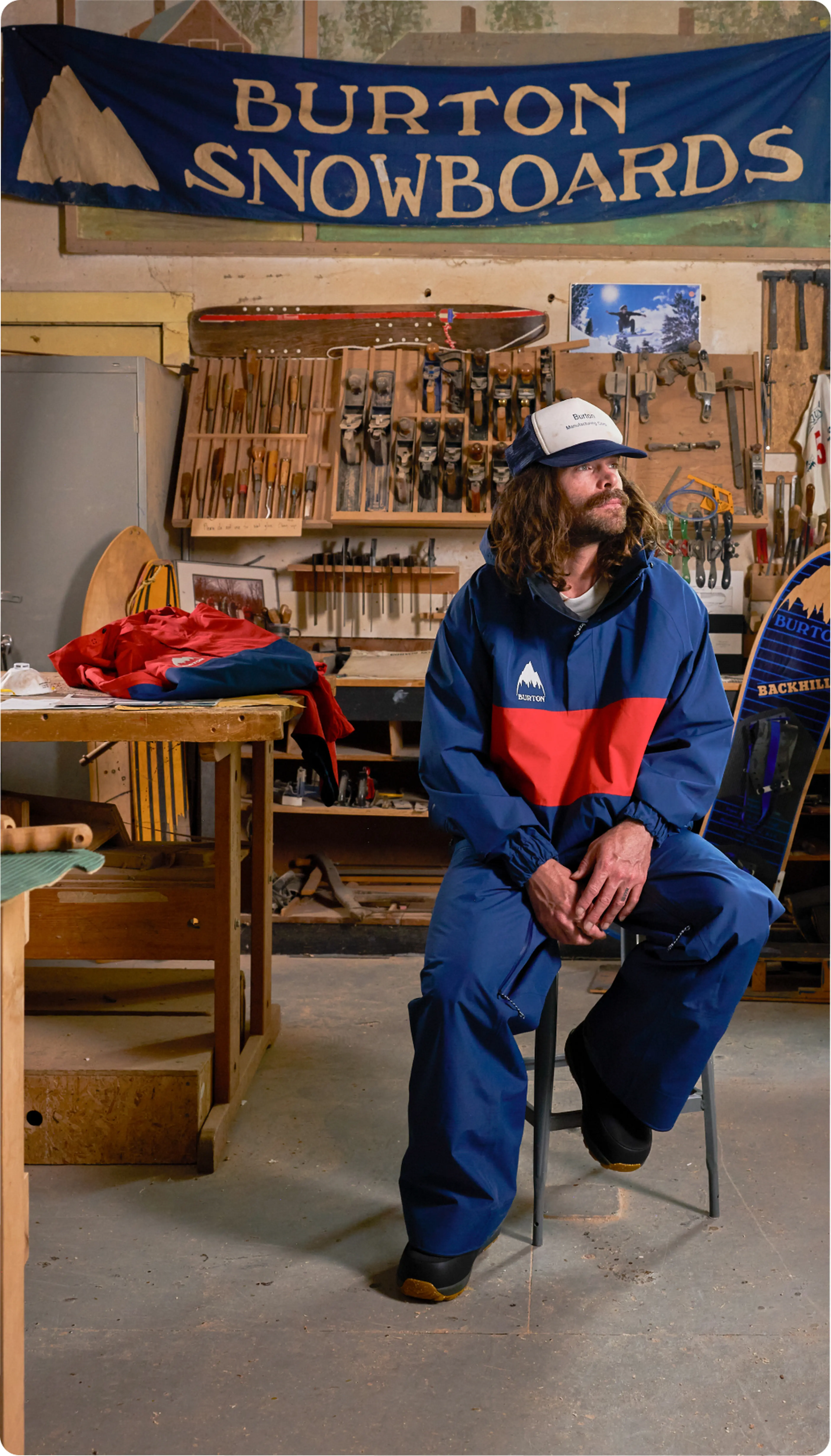 A person with long hair and a beard wearing a blue and red snowboard suit sits on a stool in a workshop, beneath a "Burton Snowboards" banner.